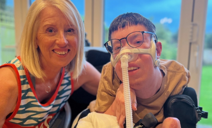 Vicky is stood next to Oli, with her arm round him. They are both smiling. Oli is wearing glasses and has a ventilator mask on. They're right in front of some patio doors. In the background you can see a bright, cloudy sky and a lawn.