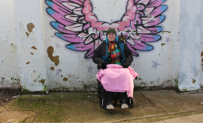 woman in a wheelchair with a pink blanket sits in front of a graffiti of some wings.