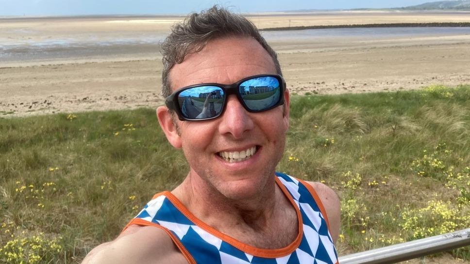 A smiling man takes a pciture by a beach in a MDUK branded sports top