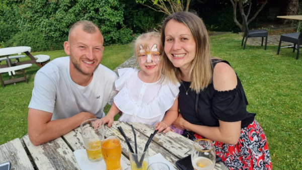 A family of three sit in a sunny pub garden. The little girl has a unicorn facepaint