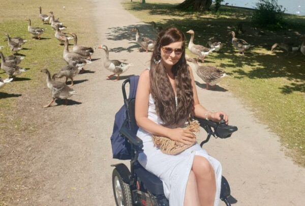a women in a long white dress sits in the sun surrounderd by geese