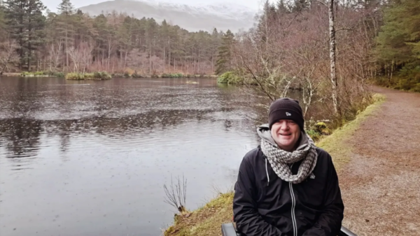 A wrapped up man sits by a beautiful lake in a forest
