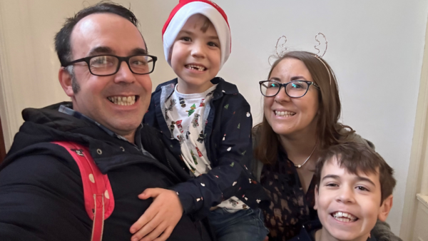 A smiling family of four pose for the camera. Two are werig christmas hats