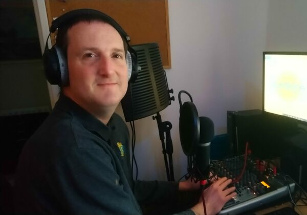 A man wearing headphones sits infront of a radio microphone and a computer