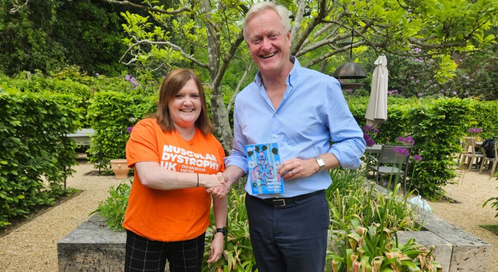 A women and a man shaking hands in a garden. The women is wearing an orange muscular dystrophy uk t-shirt.