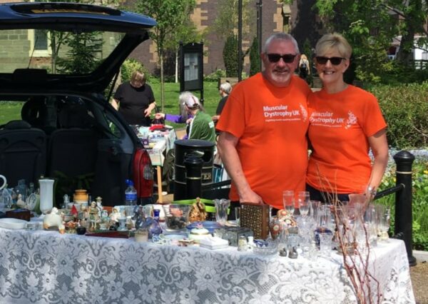 Happy couple stand by open car boot with a table full of items to sell