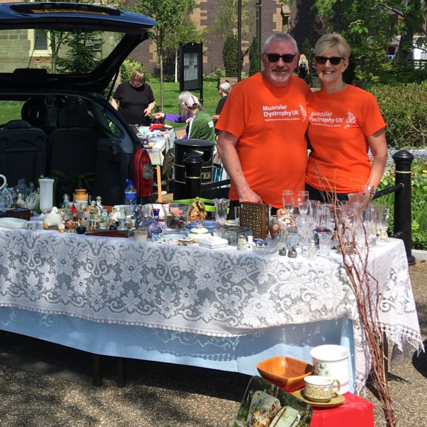 Happy couple stand by open car boot with a table full of items to sell