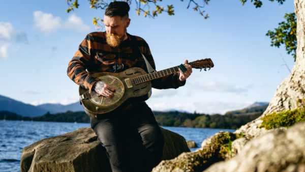 A man plays a guitar sittiing on a rock besides a large tranquil lake