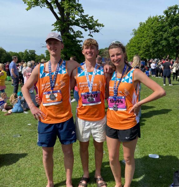 Three people pose wearing MDUK sports tops and medals on a sunny day in a field