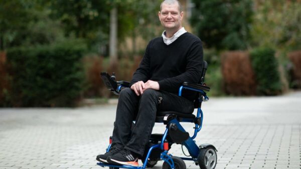 A photo of a man in a wheelchair smiling. He is wearing black clothing and black trainers