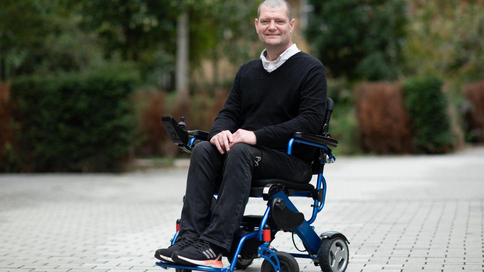A photo of a man in a wheelchair smiling. He is wearing black clothing and black trainers