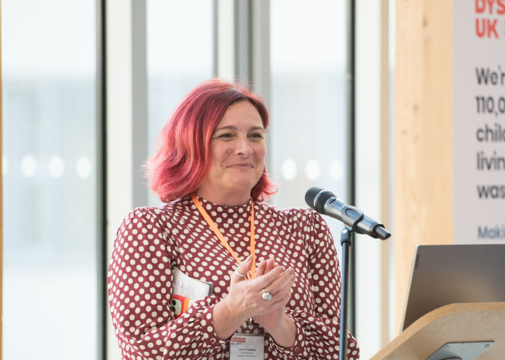 Muscular Dystrophy UK CEO, Cat Woodhead speaking. She has red hair and a pink dress on.