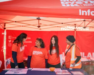 A group of volunteers stand at a desk wearing orange MDUK t-shirts.