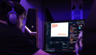 A person wearing large headphones is seated at a gaming setup, preparing for a livestream. The monitor displays a screen with the Muscular Dystrophy UK logo and the words 'Starting Soon…'. A live chat window with colourful messages is visible on the right side of the screen.