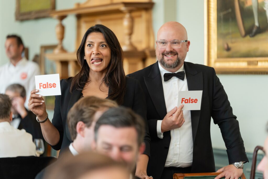 Two people in formal attire are standing and holding up cards that say "False" in bold red letters. The woman on the left is smiling playfully with her tongue slightly out, and the man on the right is smiling warmly while wearing glasses and a black tuxedo. They appear to be participating in a quiz or game event in a well-lit room with other seated guests.