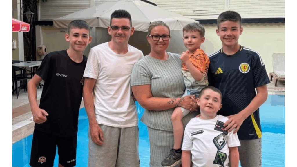 A smiling family of six stands together by a swimming pool. Two adults are in the middle with four children, two older boys on either side, a younger child in the mother’s arms, and a younger boy in front wearing a white T-shirt with graphic patches