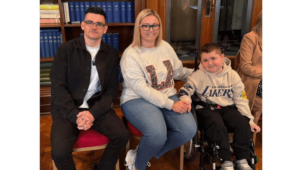 A man, woman, and young boy in a wheelchair sit side by side in front of a bookcase filled with books. The woman holds the boy’s hand, and all three are smiling. The boy is wearing a Los Angeles Lakers hoodie.