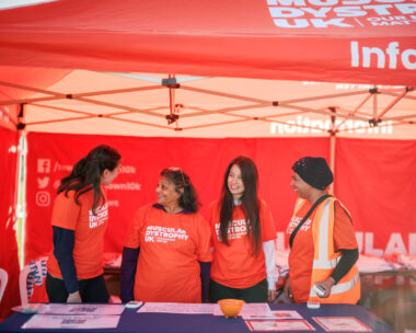 Four women at a Muscular Dystrophy UK booth under an orange tent, wearing orange shirts and smiling. One wears a hi-vis vest; info materials are on the table.