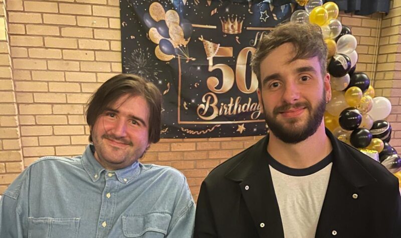 Two men sitting at a table in front of a "50th Birthday" backdrop with black, gold, and white balloons and decorations. The man on the left wears a light blue button-up shirt, while the man on the right wears a black jacket over a white shirt. A smartphone is on the table in front of them