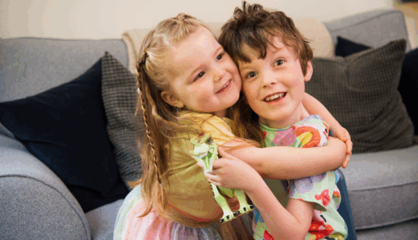 Two children sitting on a grey couch with dark cushions, hugging each other. One child wears a colorful dress and holds a toy, while the other wears a shirt with colorful patterns.