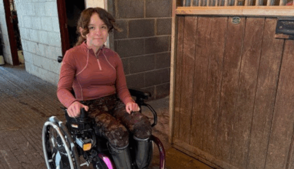 A smiling teenage girl in a wheelchair is next to a door in what appears to be a stables