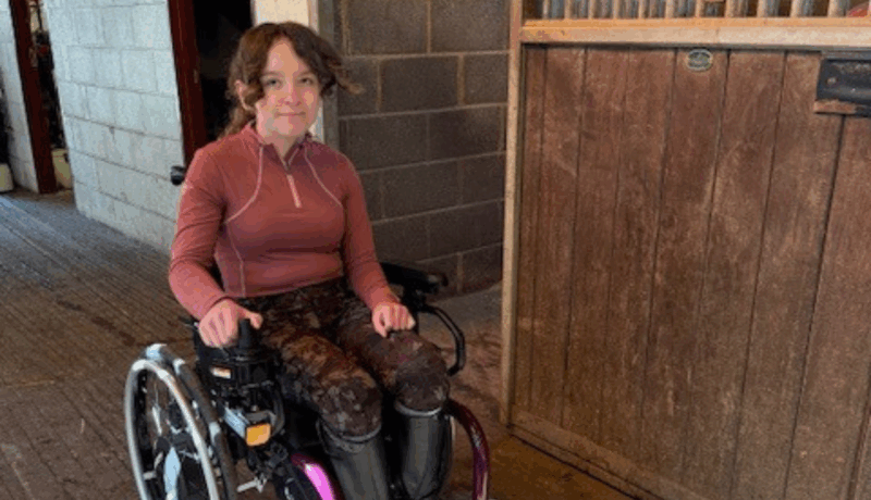 A smiling teenage girl in a wheelchair is next to a door in what appears to be a stables
