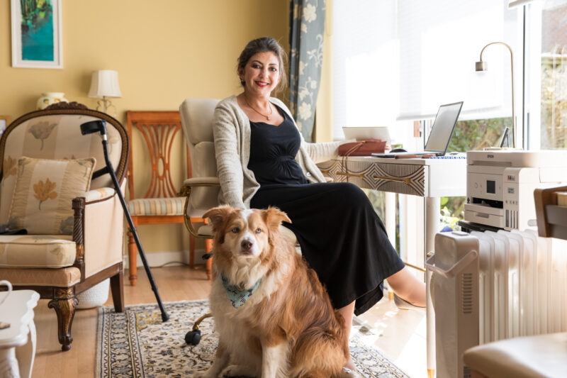 A woman sits in a living room, wearing a black dress and a light cardigan, with a walking cane nearby. A dog with a blue bandana sits at her feet. The room features furniture, a desk with a laptop and printer, and large windows letting in natural light.