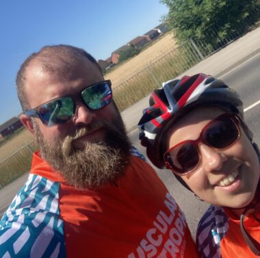 Two people take a selfie. One wears an orange cycling top, glasses and has a beard. Another has a cycling helmet on, wears glasses and an orange top.