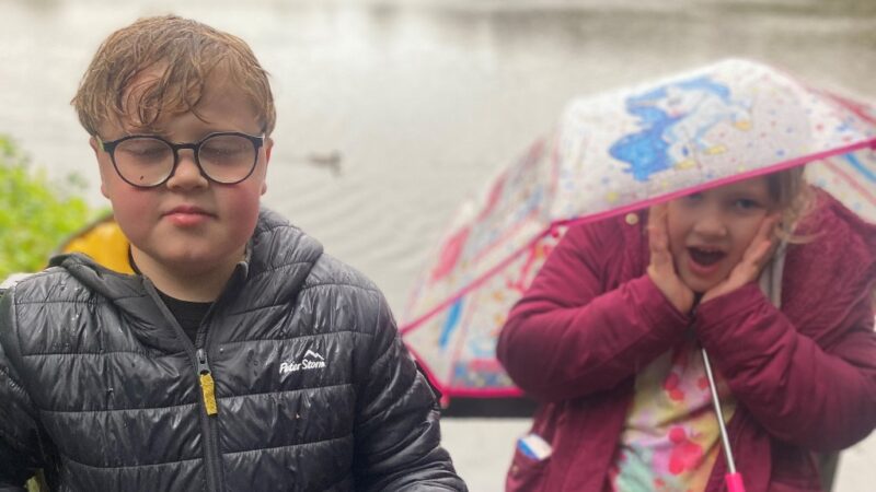 Two children outside in the rain. One wears glasses with a black jacket. The other holds a colourful umbrella and has a surprised expression, wearing a pink jacket.