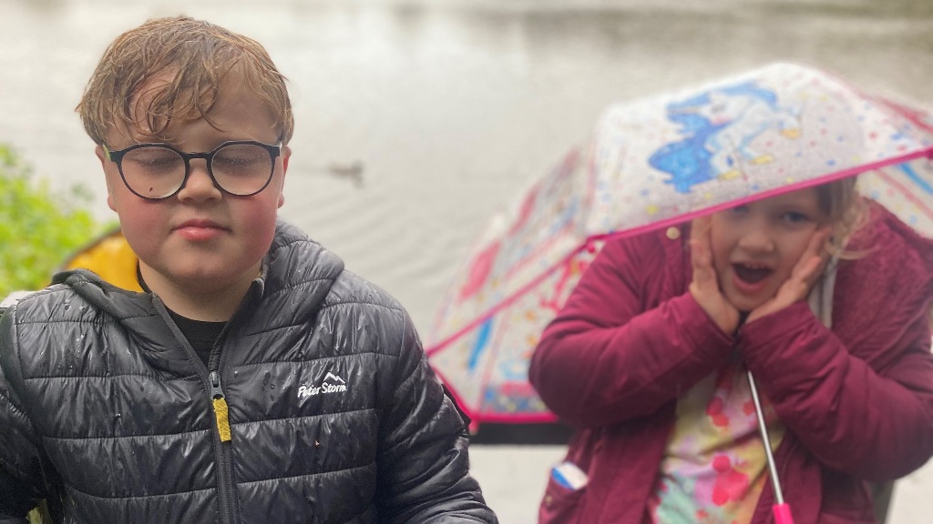 Two children outside in the rain. One wears glasses with a black jacket. The other holds a colourful umbrella and has a surprised expression, wearing a pink jacket.