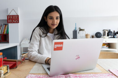 Person sitting at a table using a laptop with 'Muscular Dystrophy UK' sticker, in a cosy room with shelves, jars, and a 'FREE HUGS' sign on the wall.