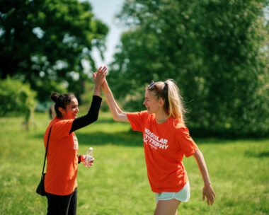 Two people in matching orange 'Muscular Dystrophy UK' T-shirts high-five in a park, one holding a water bottle and wearing a black shoulder bag.