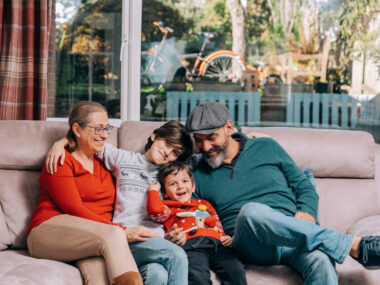 Family of four sitting together on a couch. Adults wear red and blue tops, children wear patterned grey and festive red sweaters. A large window behind them reveals greenery and a bicycle.
