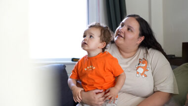 An adult and a child sitting together on a sofa. The adult wears a light shirt with a cartoon dog illustration from Bluey, and the child wears an orange shirt with the word 'Happy'.