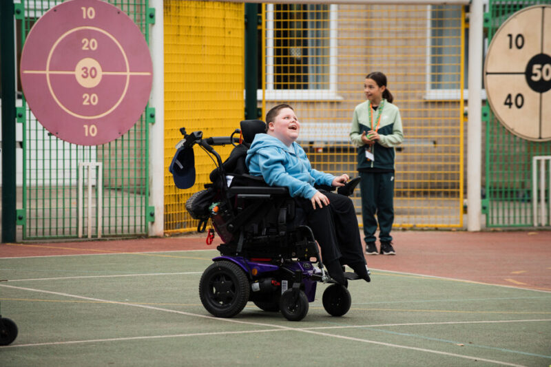 A person in a motorised wheelchair on an outdoor sports court, accompanied by another standing individual. In the background, two large target boards display concentric circles with numbers ranging from 10 to 50.
