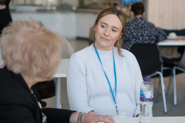 A woman smiles while looking at another person sat at a table
