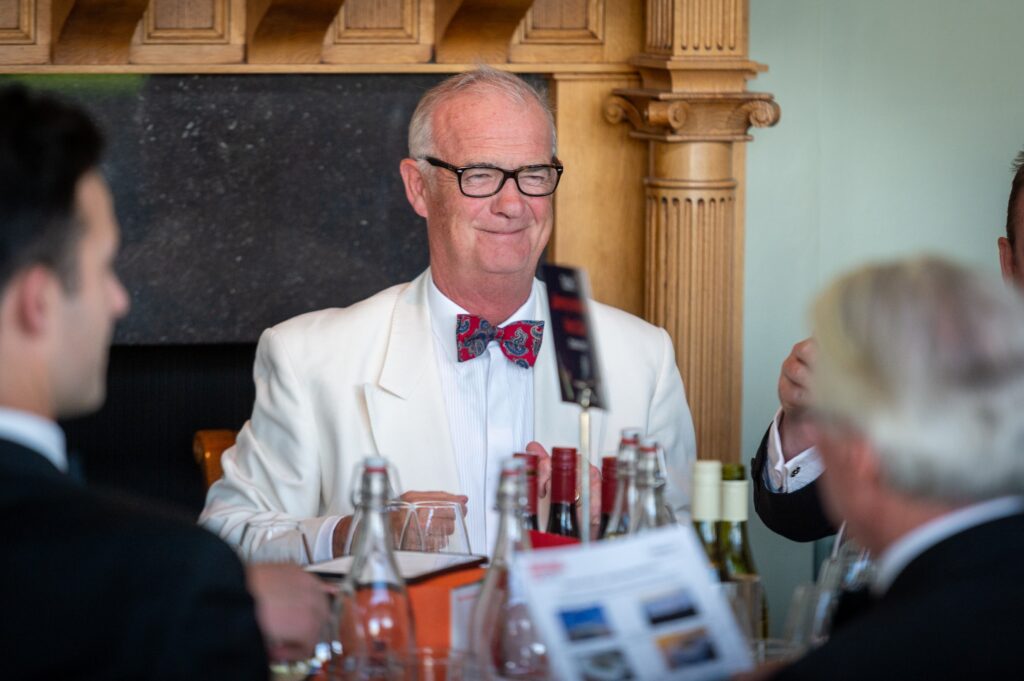 A person in a white suit with glasses on sits at a table with others.