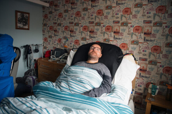 A person with glasses on and a grey jumper lies in bed looking upwards.