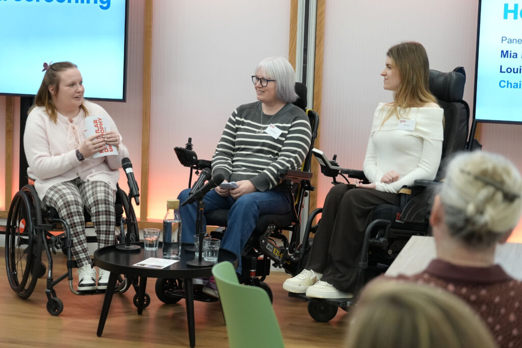 Three people in wheelchairs are participating in a panel discussion. They are in front of a large screen. A small round table in front of them holds water bottles, glasses, and printed materials.