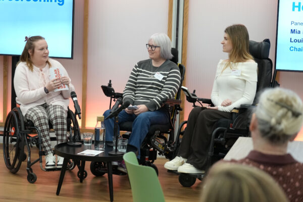 Three people in wheelchairs are participating in a panel discussion. They are in front of a large screen. A small round table in front of them holds water bottles, glasses, and printed materials.