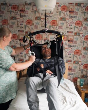 A person with glasses is being lifted in a sling above a bed with another person standing beside them adjusting the straps. The room has patterned wallpaper and a bedside table.