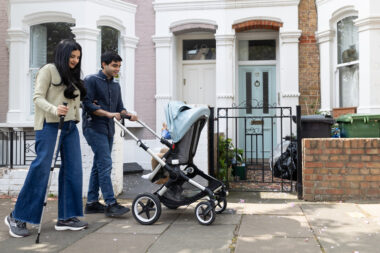 A woman using a mobility stick walks arm-in-arm with a man who is pushing a pushchair with a child in. They are walking along a pavement in front of a row of terraced houses.