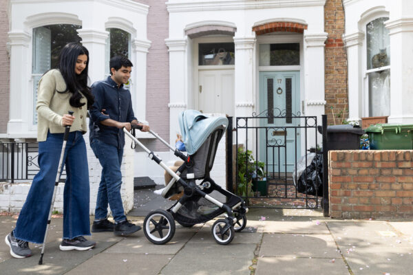 A woman using a mobility stick walks arm-in-arm with a man who is pushing a pushchair with a child in. They are walking along a pavement in front of a row of terraced houses.