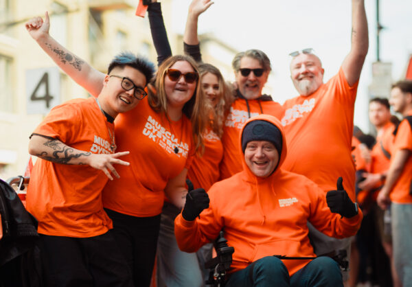 Group of people in bright orange “Muscular Dystrophy UK” T‑shirts posing on a street, at what looks like a charity run, with one person in a powered wheelchair at the front giving thumbs up and others raising their arms.