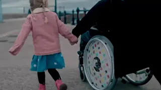 Video thumbnail showing a child walking beside a wheelchair user on a waterfront promenade, holding hands as they move along a paved path.