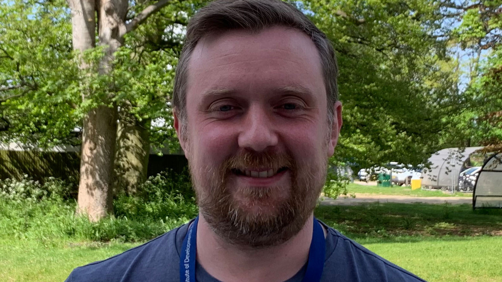 Headshot of Tom Roberts, the background appears to be a park with lots of grass and trees. You can also see the top of Tom's t-shirt which is navy in colour and his blue lanyard.