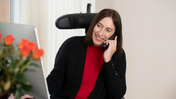 A woman who has FSHD and uses a wheelchair smiles while on the phone as she sits in front of a computer screen.