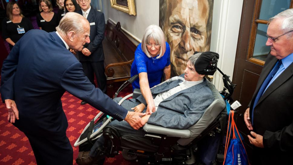 Duke of Edinburgh shakes hands with a man in a powerchair at an MDUK event while other attendees watch on.