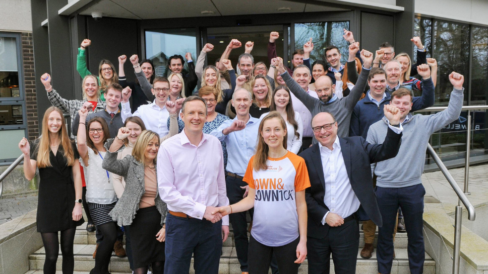 Staff from Bidwells head office are all stood in front of the building with one arm in the, cheering. At the front is Matt Allen and Amy O'Hanlon shaking hands