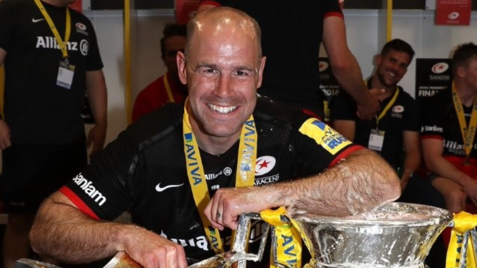 Charlie Hodgson smiling with a medal round his neck, leaning on the Aviva Premiership trophy